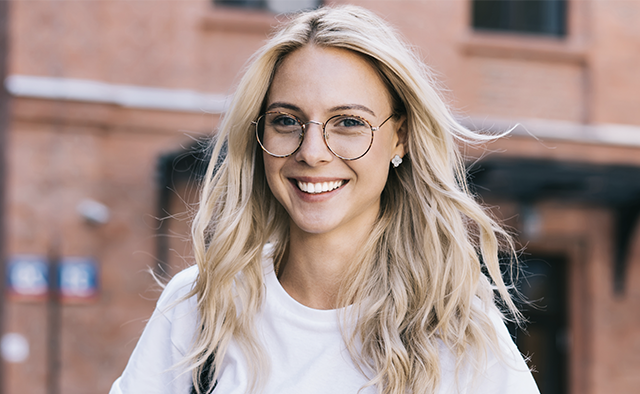 Photo: Smiling woman wearing eyeglasses and white t-shirt with brick building in the background