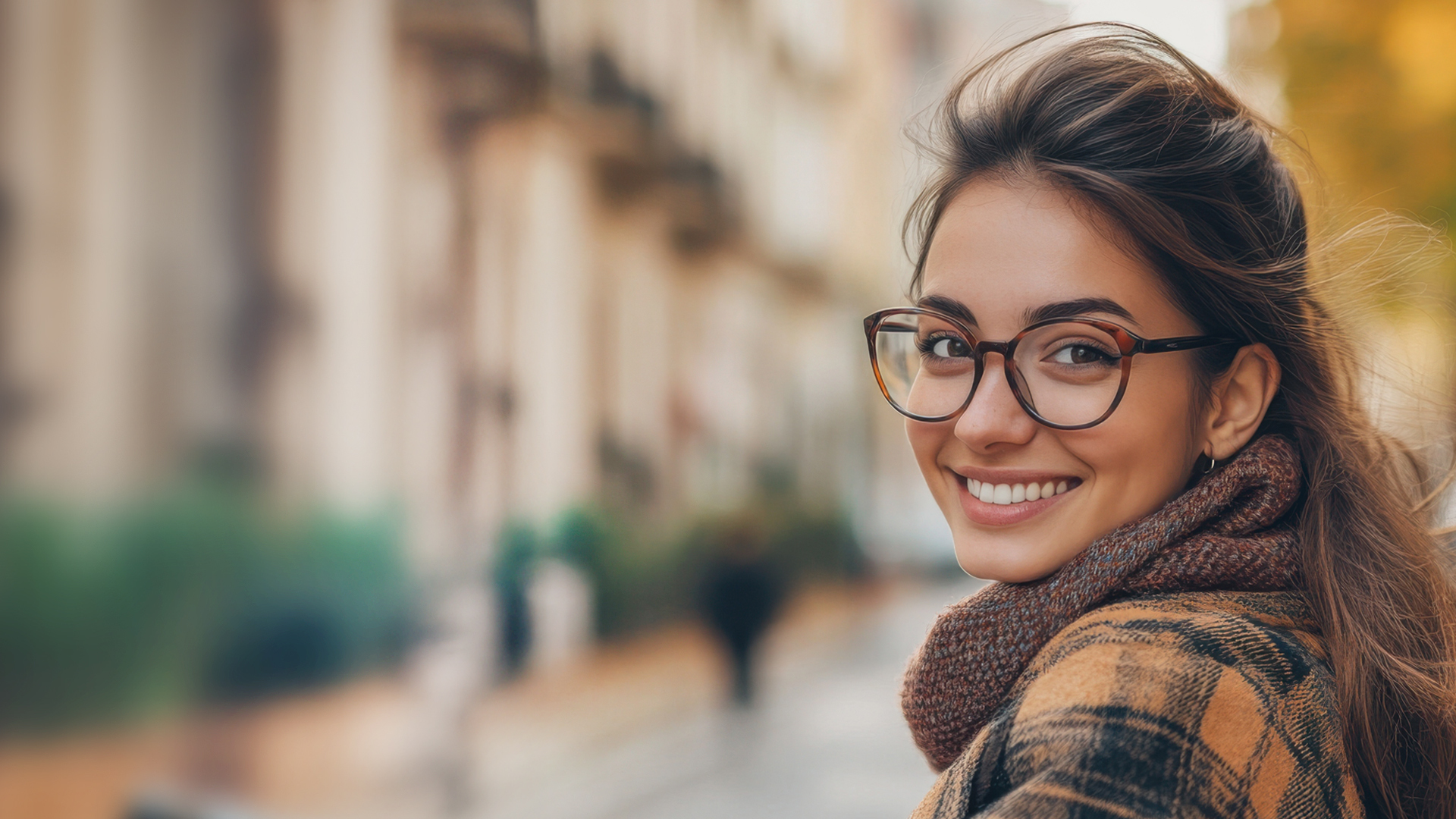 Girl with long hair and glasses smiling over her shoulder, wearing a plaid coat and knitted scarf in an autumn setting with yellow-leaved trees and blurred buildings in the background