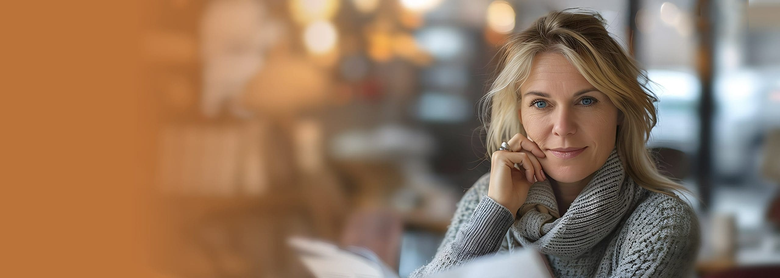 A woman wearing a knit scarf is seated, holding and reading a book. The background is softly blurred with warm ambient lighting.