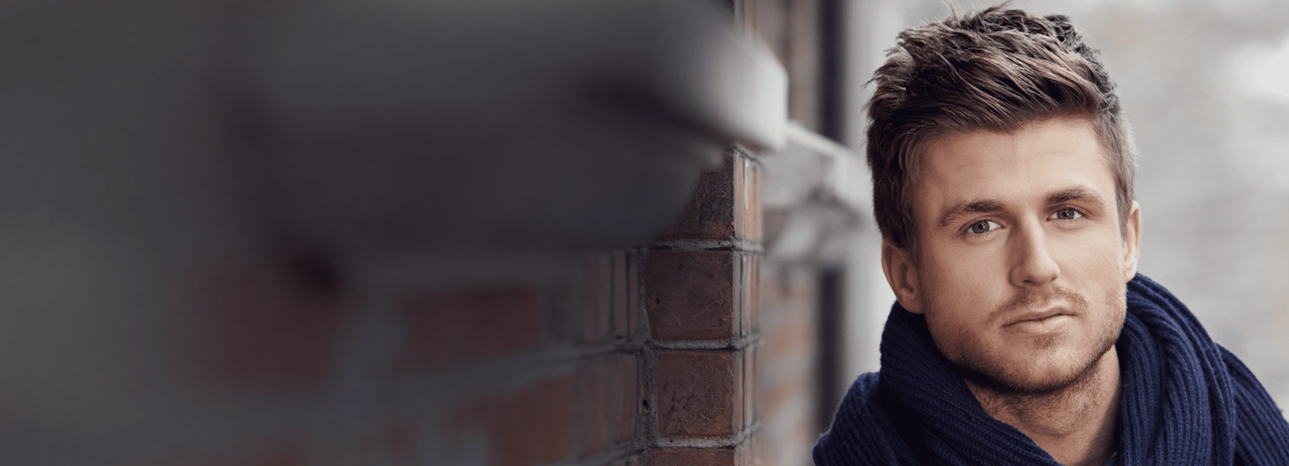 A man with short, styled hair wearing a dark scarf, leaning against a brick wall. The background is blurred.