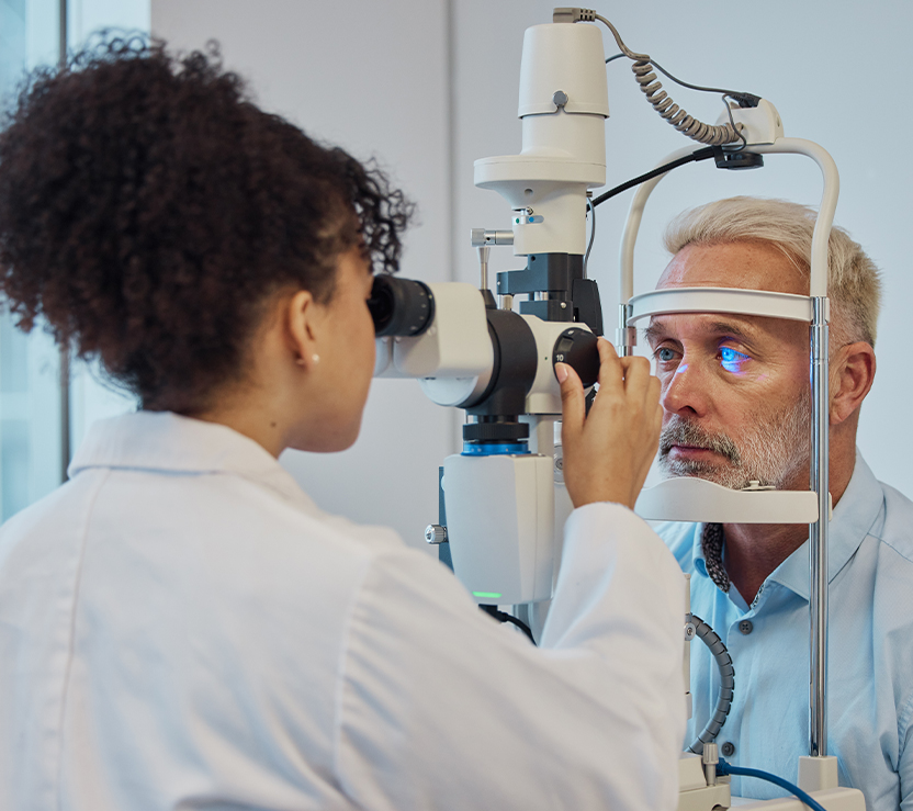 An eye care professional conducting an eye exam. The patient is seated with their chin on the support while the professional adjusts the equipment.